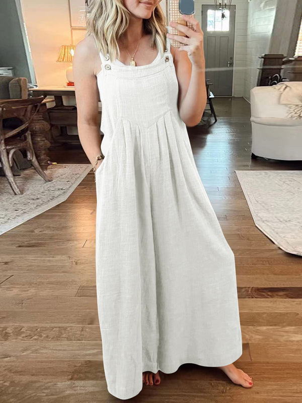 A woman wearing a white linen wide-leg overall jumpsuit with a relaxed fit, sleeveless top, and open-toe sandals, standing in a room with wooden flooring.